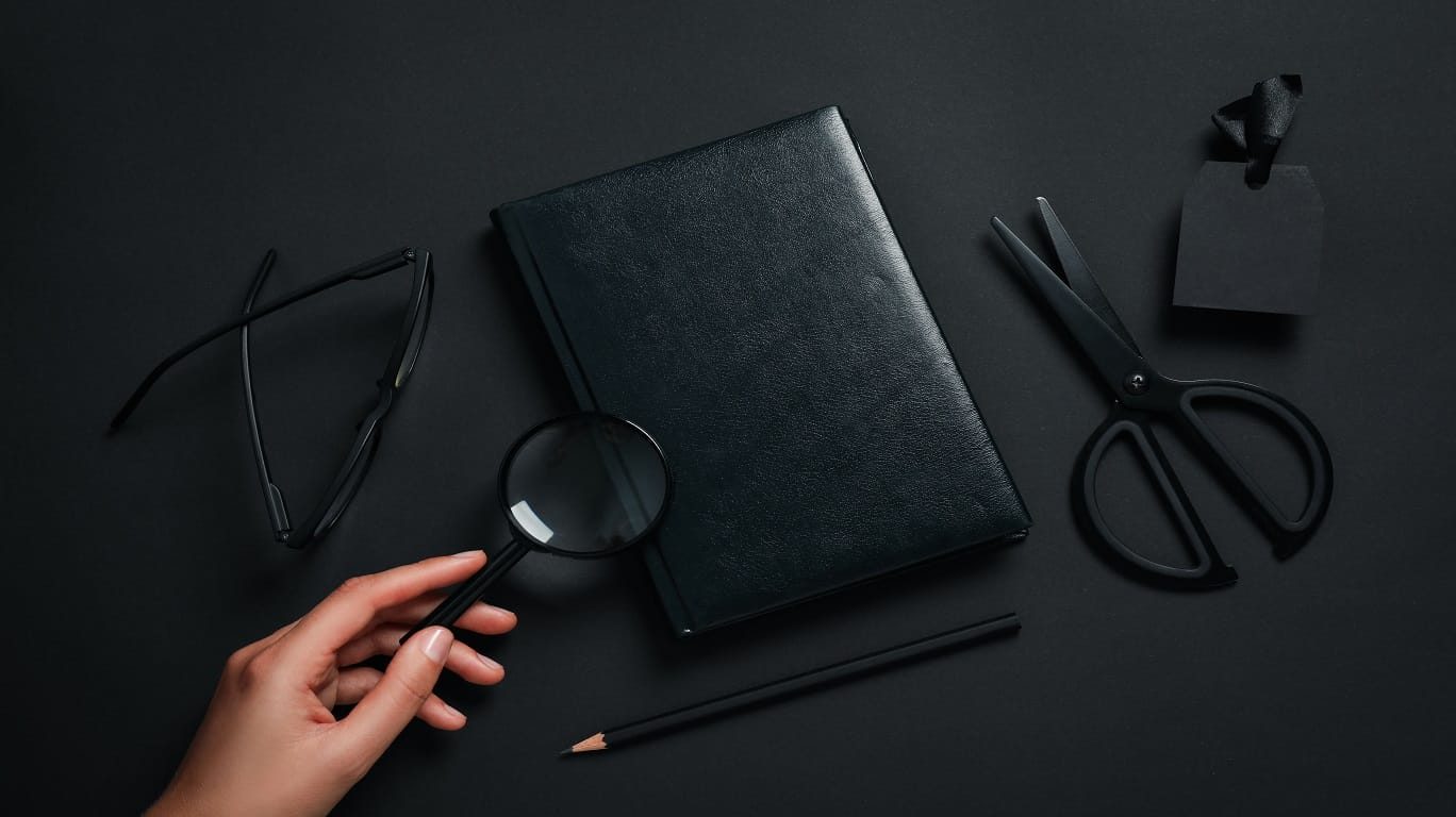Hand holding a magnifying glass over a black book cover, with black glasses, scissors, pencil and a blank tag on a dark background
